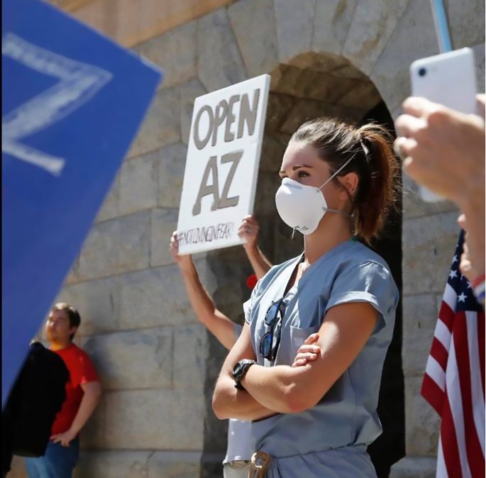 En ensam ung kvinna med korslagda armar i munskydd står bland protesterande människor plakat, helt lugn i röran